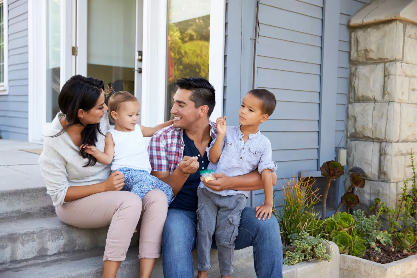  Una madre y un padre sentados en el porche de su casa con sus dos hijos pequeñosndo
