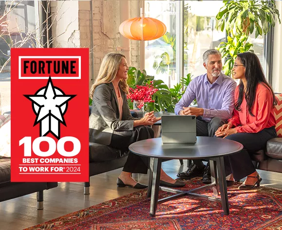  Three colleagues sitting on couches in a modern, brightly lit office space, having a discussion around a coffee table with a laptop on it. Next to them is a sign that reads 'Fortune 100 Best Companies to Work For 2024.
